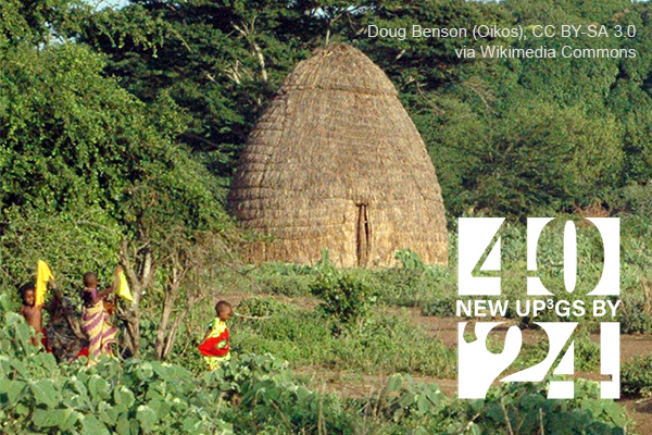 people walk by a thatched hut in a wooded area. Doug Benson (Oikos) CC BY-SA 3.0 via Wikimedia Commons.