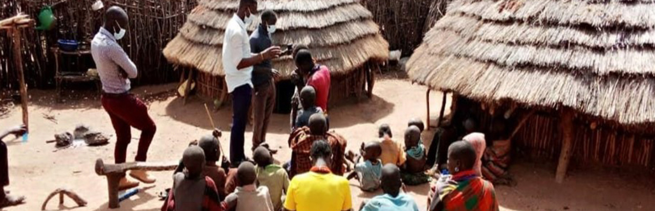 a group of people sit outside of a thatched hut and listen to a teaching